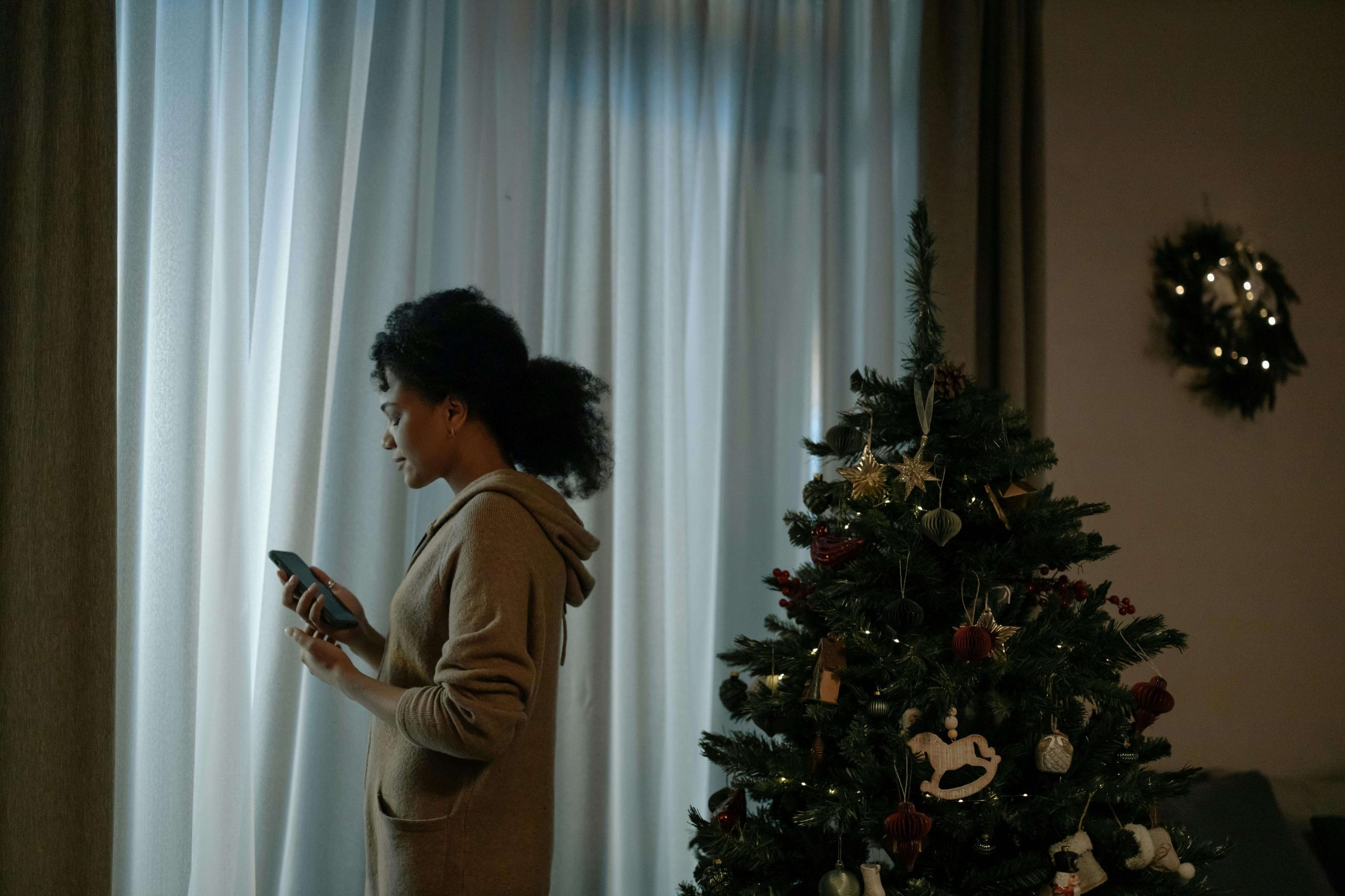 Woman standing by a window near a Christmas tree, checking her phone while coping with holiday overload, illustrating support from a psychiatrist for stress in Tempe, AZ.