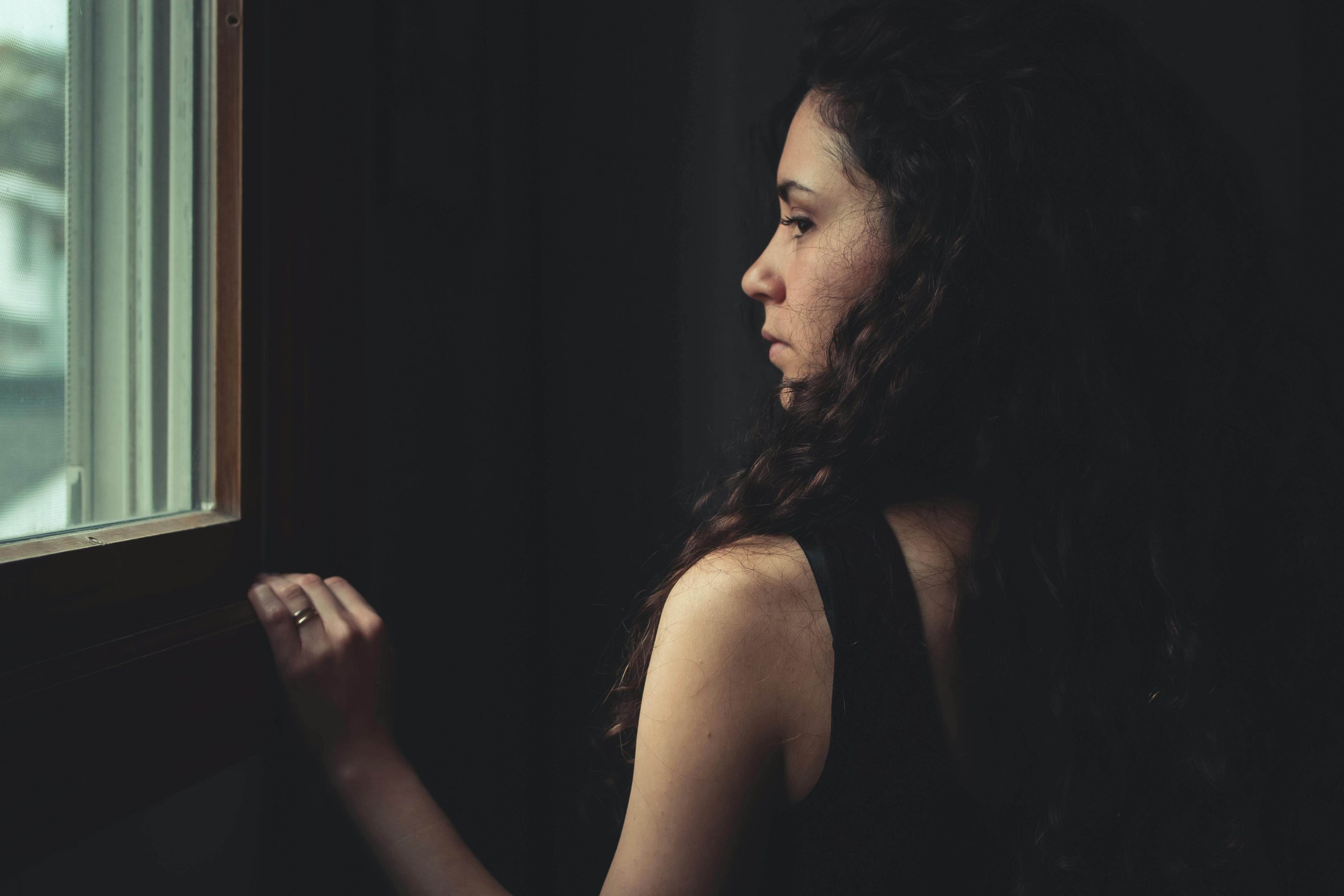Woman Standing by Window - iop for substance use tempe, az - iop substance abuse treatment - intensive outpatient programs - phx - phoenix - gilbert - scottsdale - mesa - arizona - chandler Woman standing by a window in dim light, reflecting on her emotions—representing the introspection and healing that begins through IOP for substance use in Tempe, AZ.