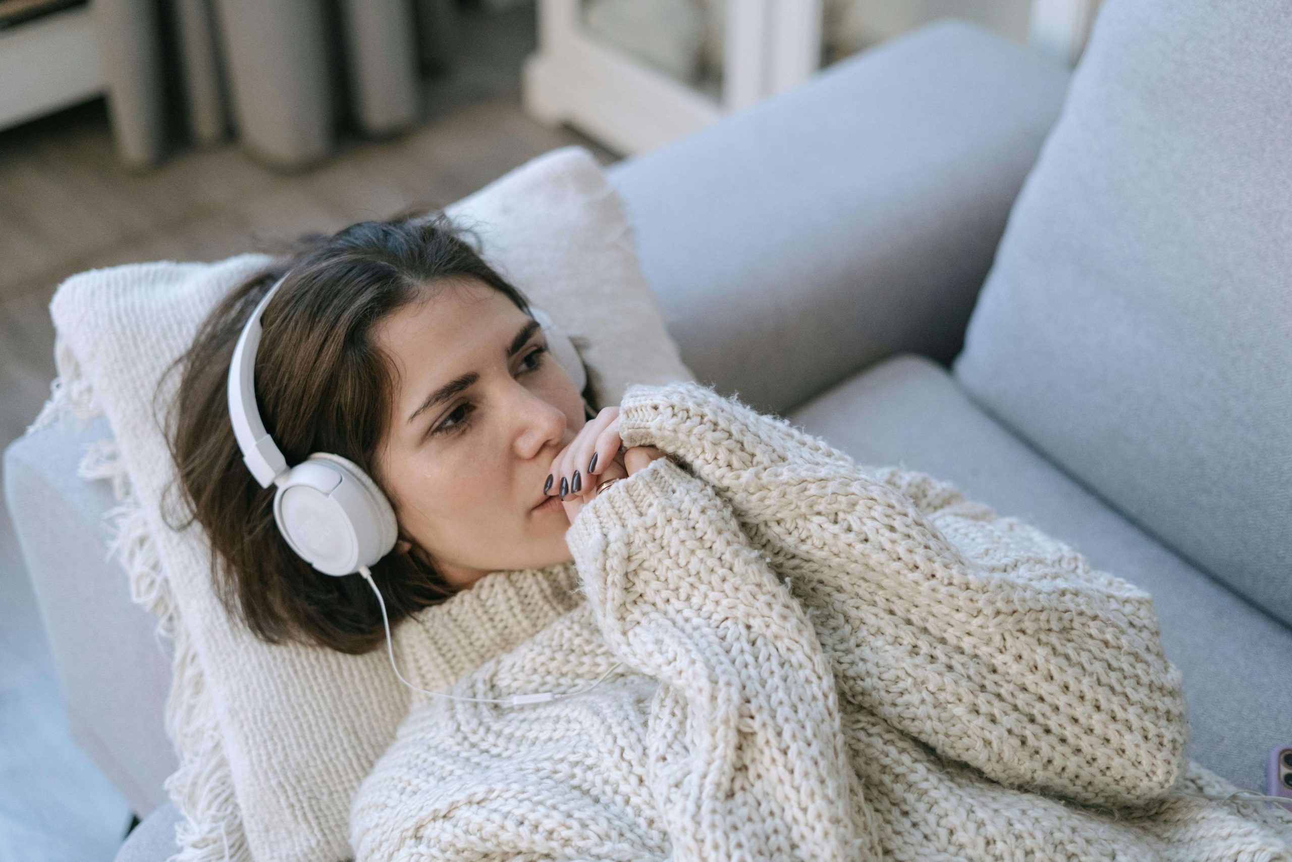 A woman in a cozy sweater lying on a couch with headphones on, listening thoughtfully, illustrating relaxation techniques and self-care practices used in bipolar disorder therapy in Tempe AZ.
