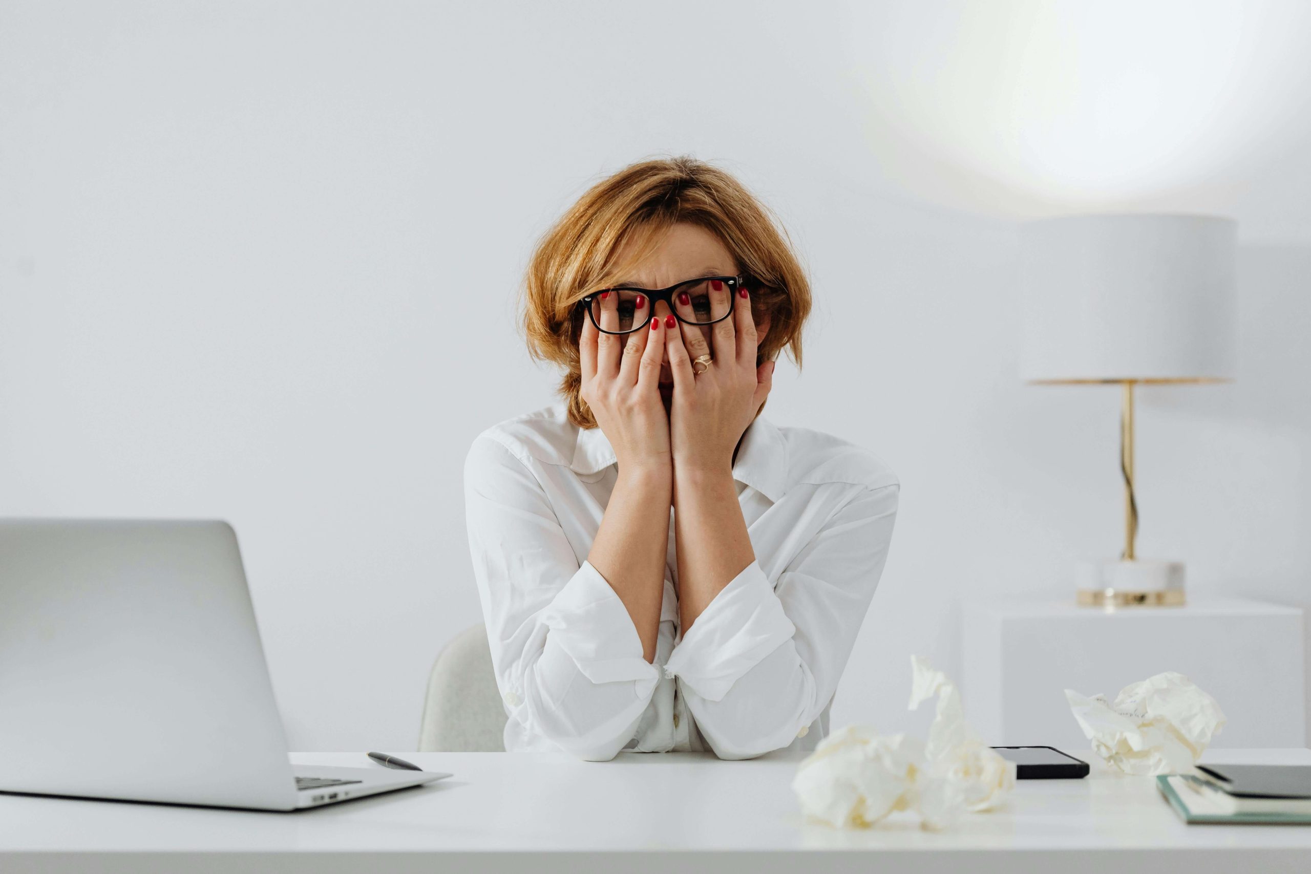 A woman covers her face in frustration while sitting at a white desk with crumpled papers and a laptop, capturing the emotional strain ADHD can bring. Support is available through psychiatric care in Tempe, AZ.
