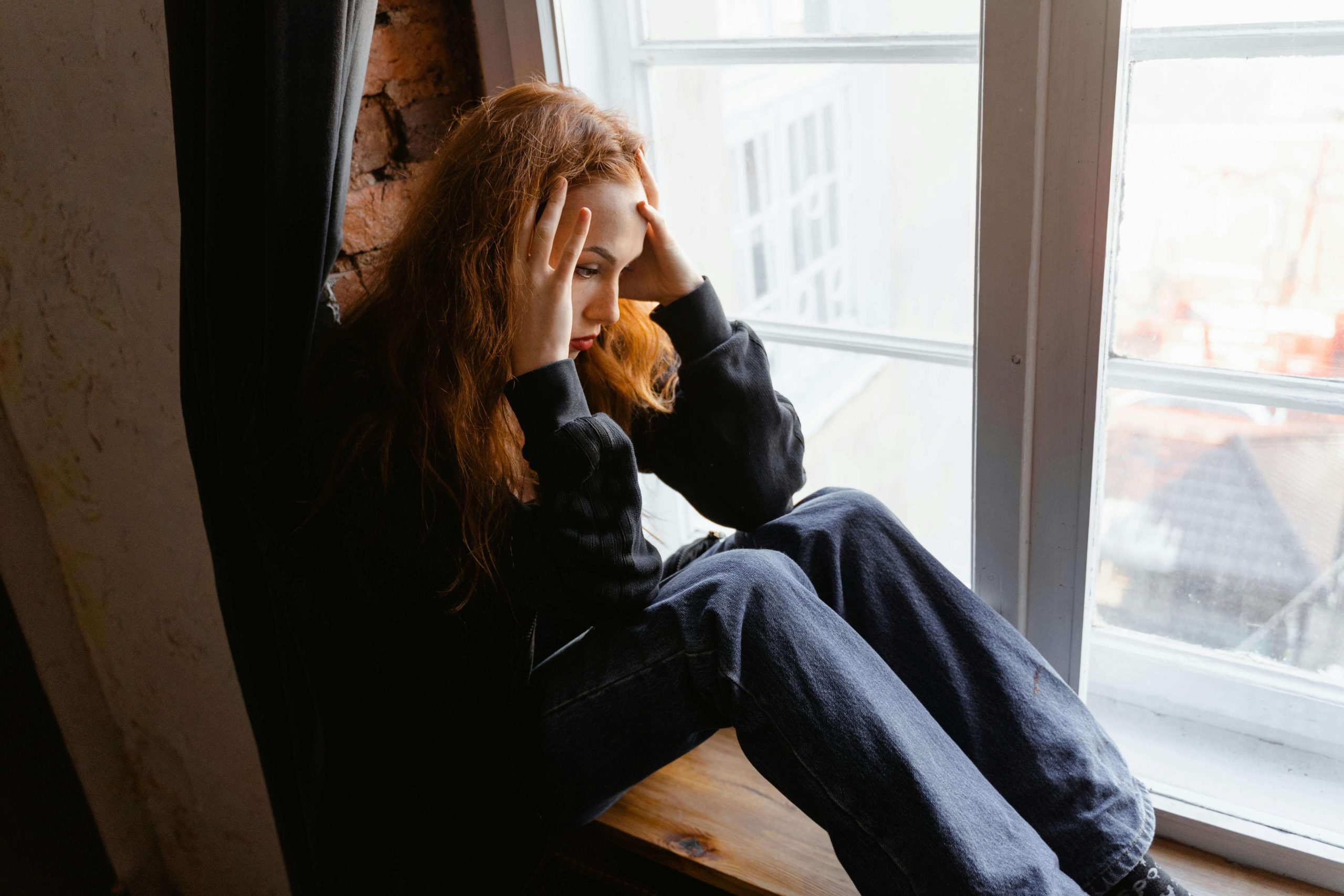 Person sitting by a window appearing deep in thought, illustrating the struggle to differentiate everyday stress from deeper mental health concerns.