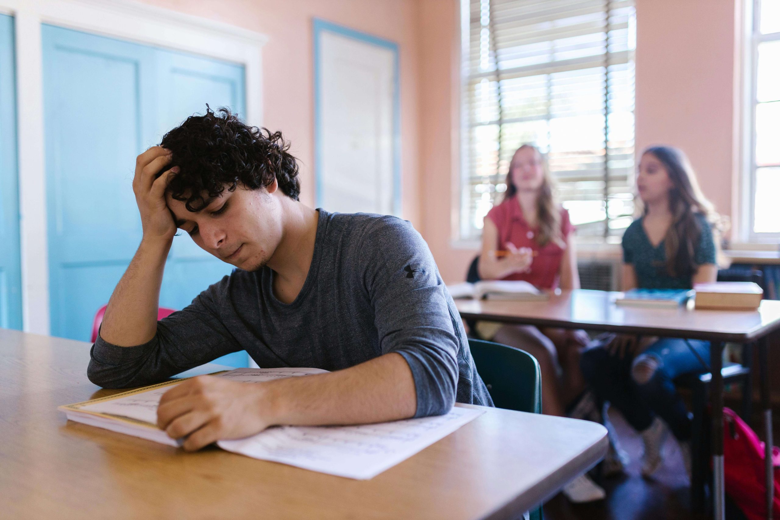 A teen boy sits with his head in his hand, struggling to focus during class while classmates talk nearby, illustrating the academic and social toll of untreated ADHD, and the relief possible through medication management in Tempe, AZ.