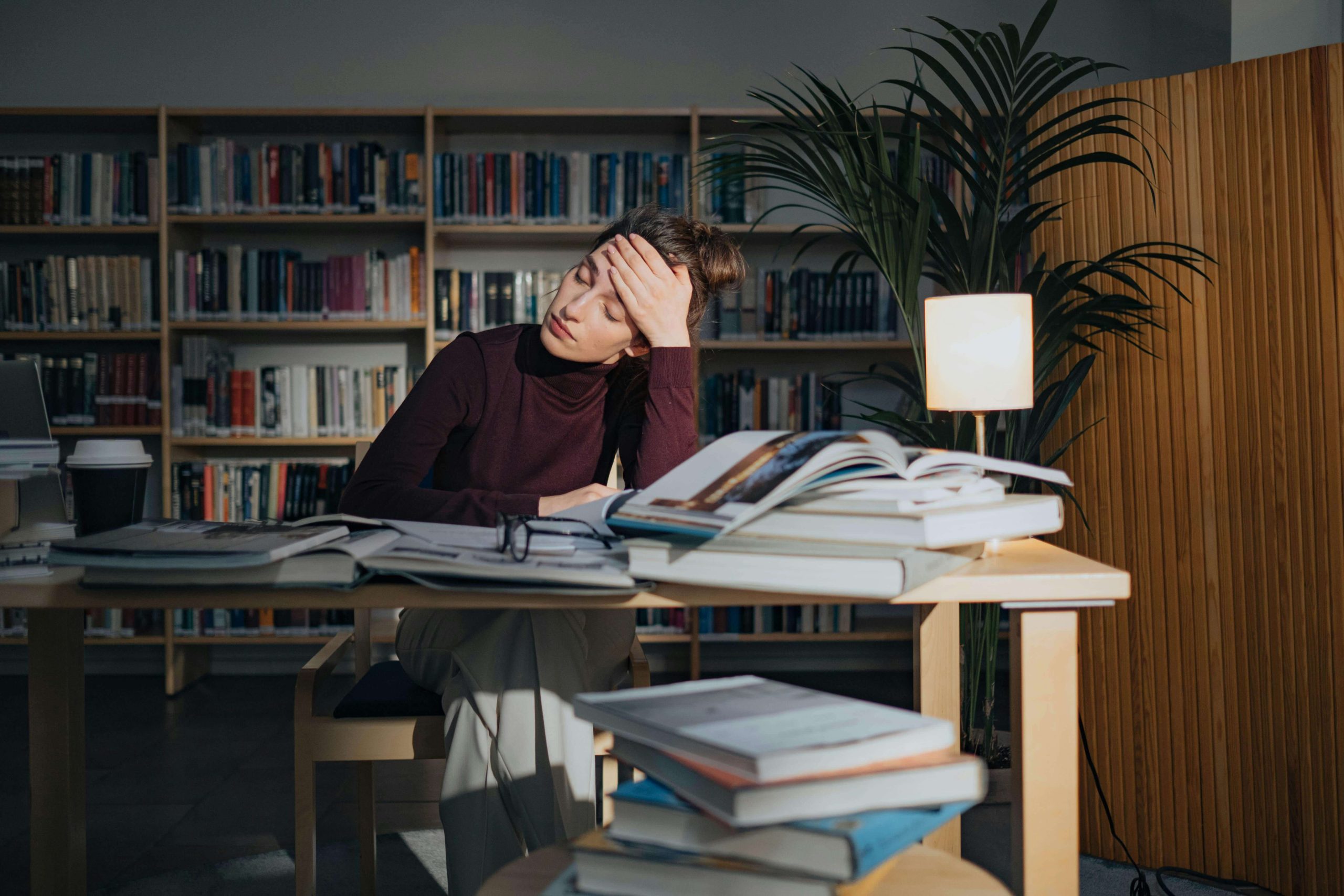 A woman sits at a cluttered library table, looking overwhelmed and holding her head in her hand, symbolizing the mental load that may lead someone to seek psychiatric care in Tempe, AZ, for adult ADHD.
