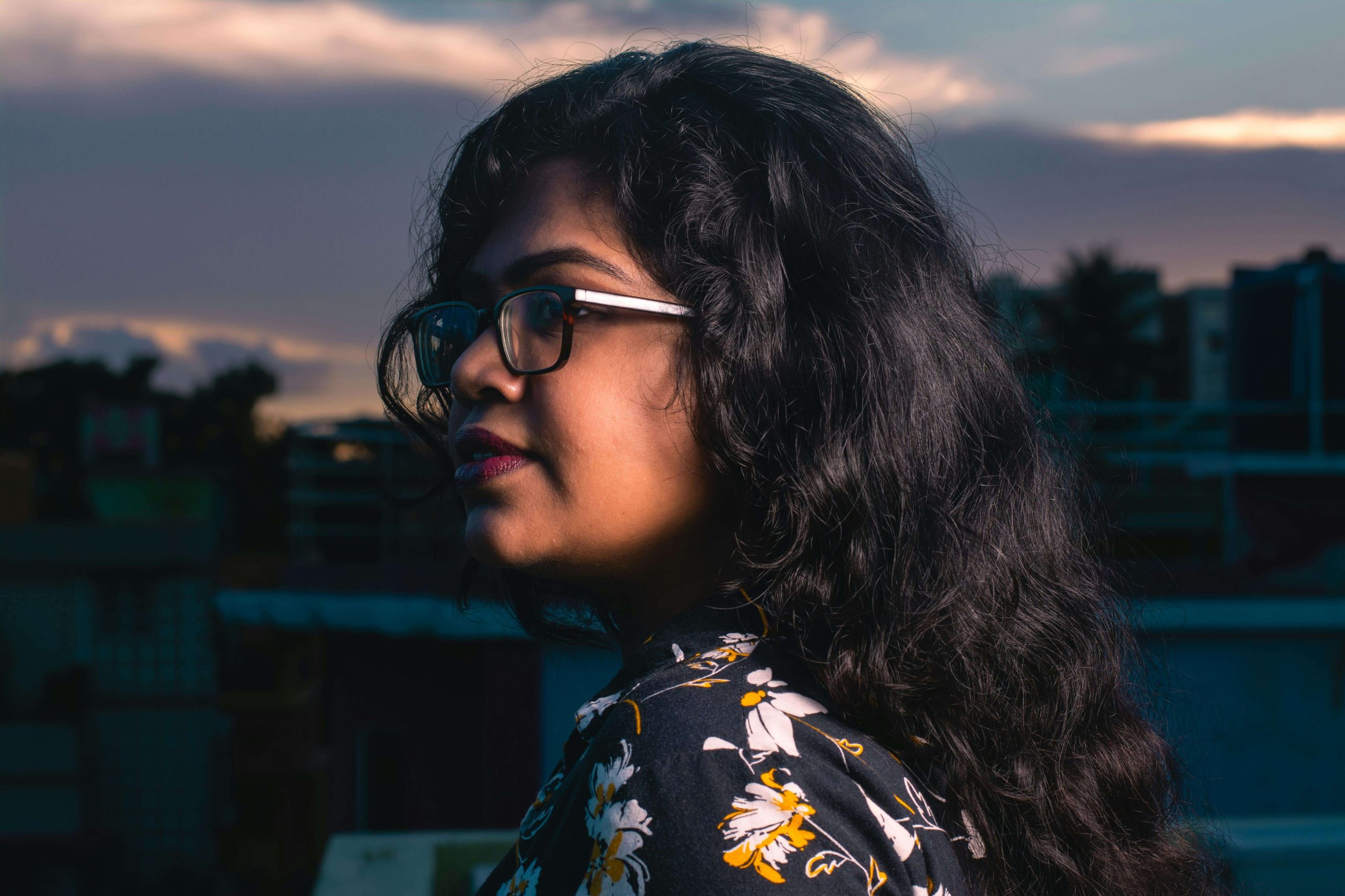 Woman Reflecting - psychiatric care tempe - psychiatry tempe az - psychiatrist tempe az - phx - phoenix - gilbert - scottsdale - mesa - arizona South Asian woman wearing glasses and a floral shirt gazing thoughtfully into the distance during sunset, symbolizing quiet strength and introspection supported through compassionate psychiatric care in Tempe, AZ.